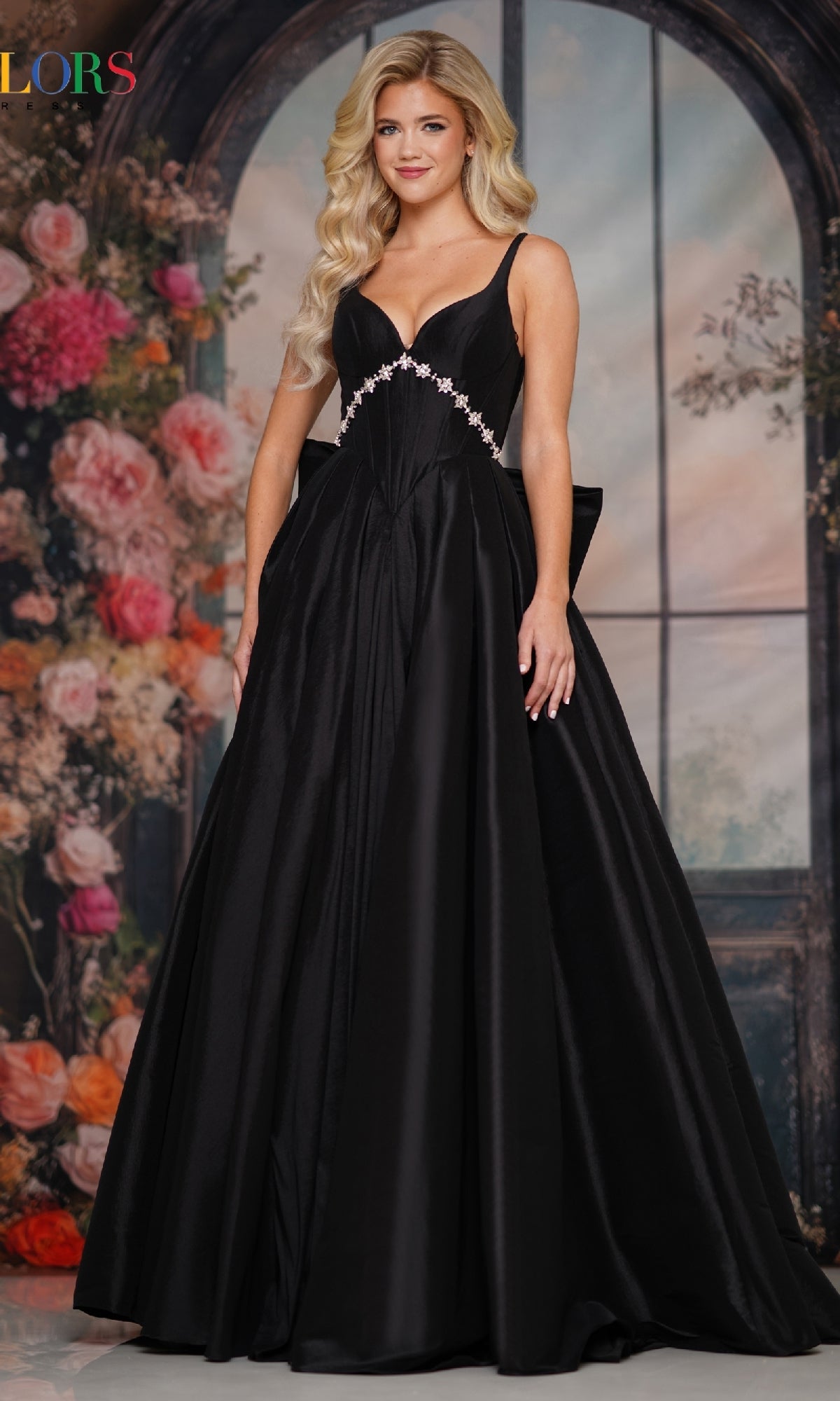 A woman wearing the Colors Dress Statement-Bow Long Prom Dress (Style 3922) stands before a floral, arched window backdrop, her long blonde waves framing a gentle smile.